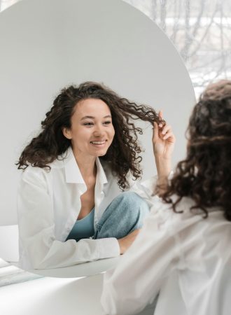 pexels-photo-6417911-6417911 Happy woman with curly hair smiling at her reflection in a mirror, embracing natural beauty.